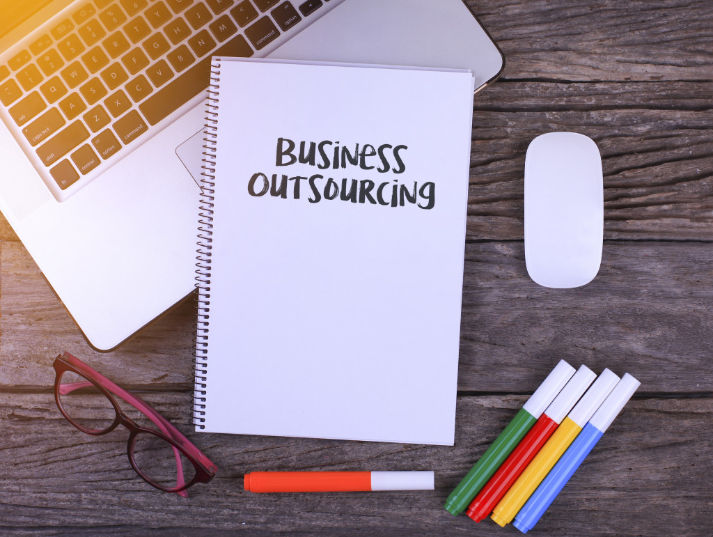 A notebook titled Business Outsourcing on an office table with a laptop, glasses, and some markers