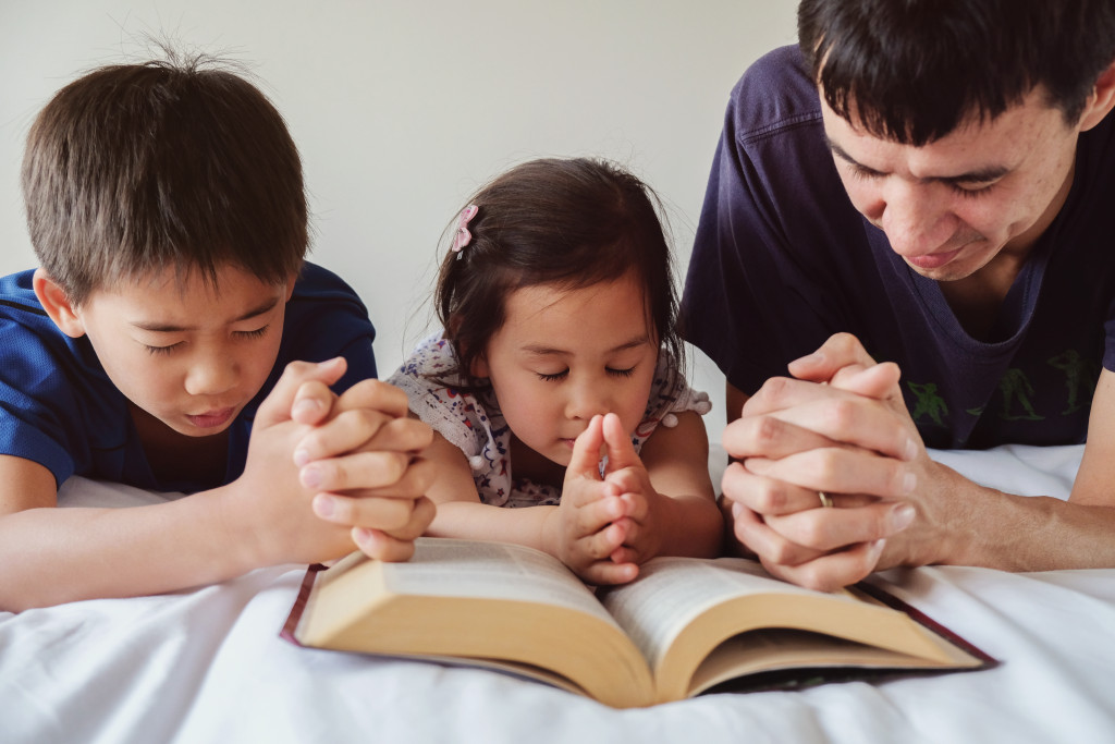 Father praying with his son and daughter while kneeling at the side of a bed with an open Bible.