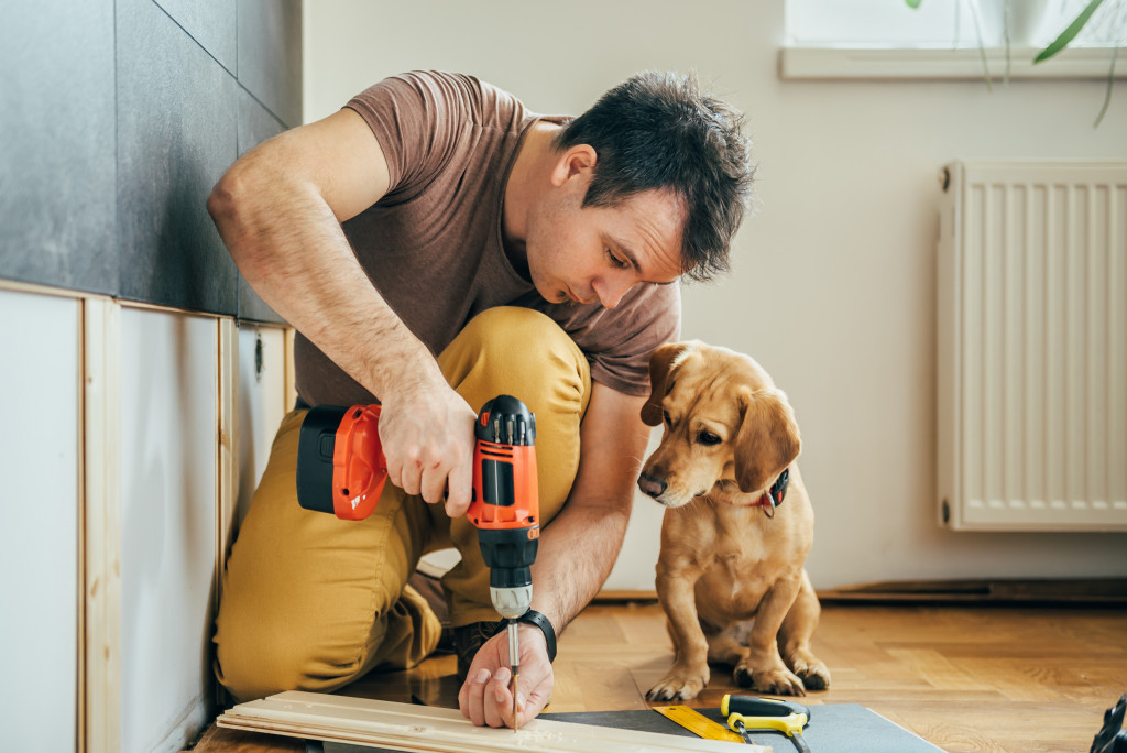 A man doing repairs with drill machine while his dog watches 