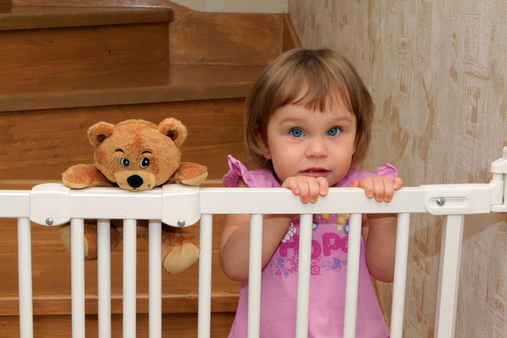 baby girl holding the baby gate with teddy bear on her side